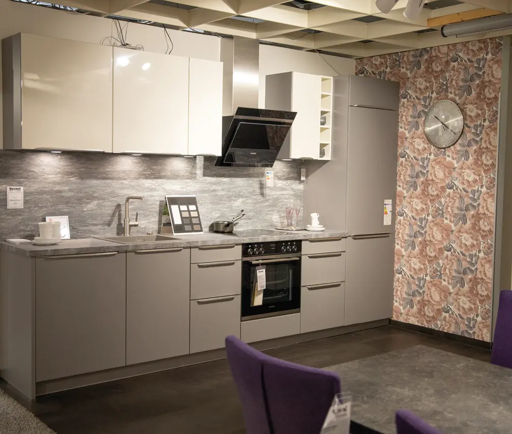 Küchenzeile mit grauen Unterschränken, weißen Hängeschränken, Spüle und Herd; rechts hoher Kühlschrank vor floraler Wand